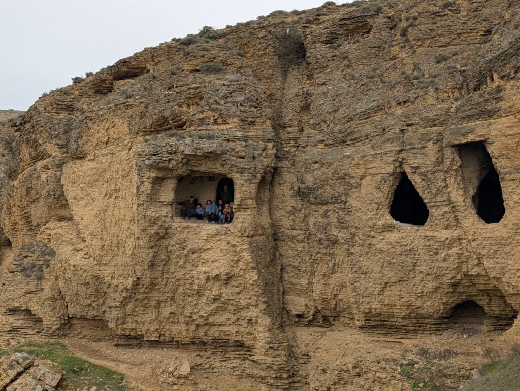 Old cave dwellings carved into a sandstone cliff at Narimankend, with visitors looking out of one of the openings