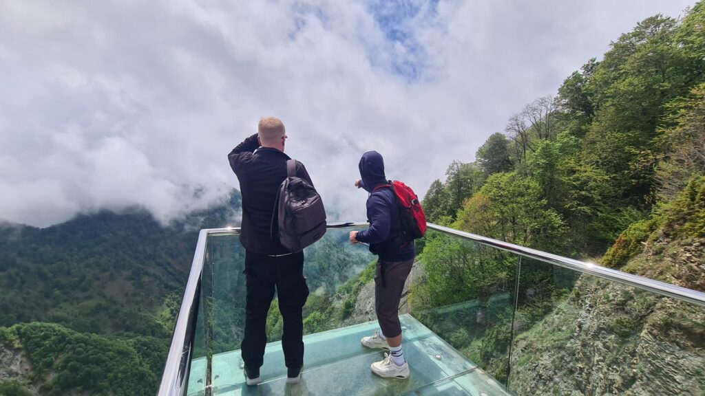 Two travellers on the glass viewing platform at Tufandag resort, above forested mountains and clouds