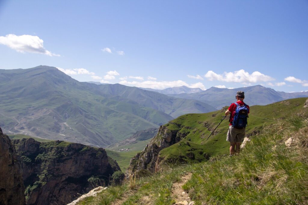 A hiker on a grassy ridge trail above a canyon, with layers of green mountains behind