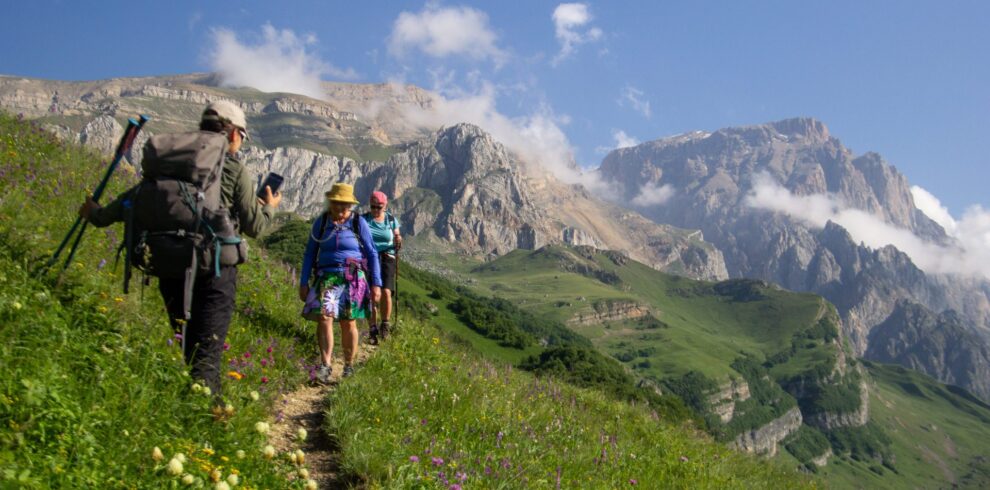 Three hikers on a narrow trail through a meadow full of wildflowers, with the limestone cliffs of Shahdag behind