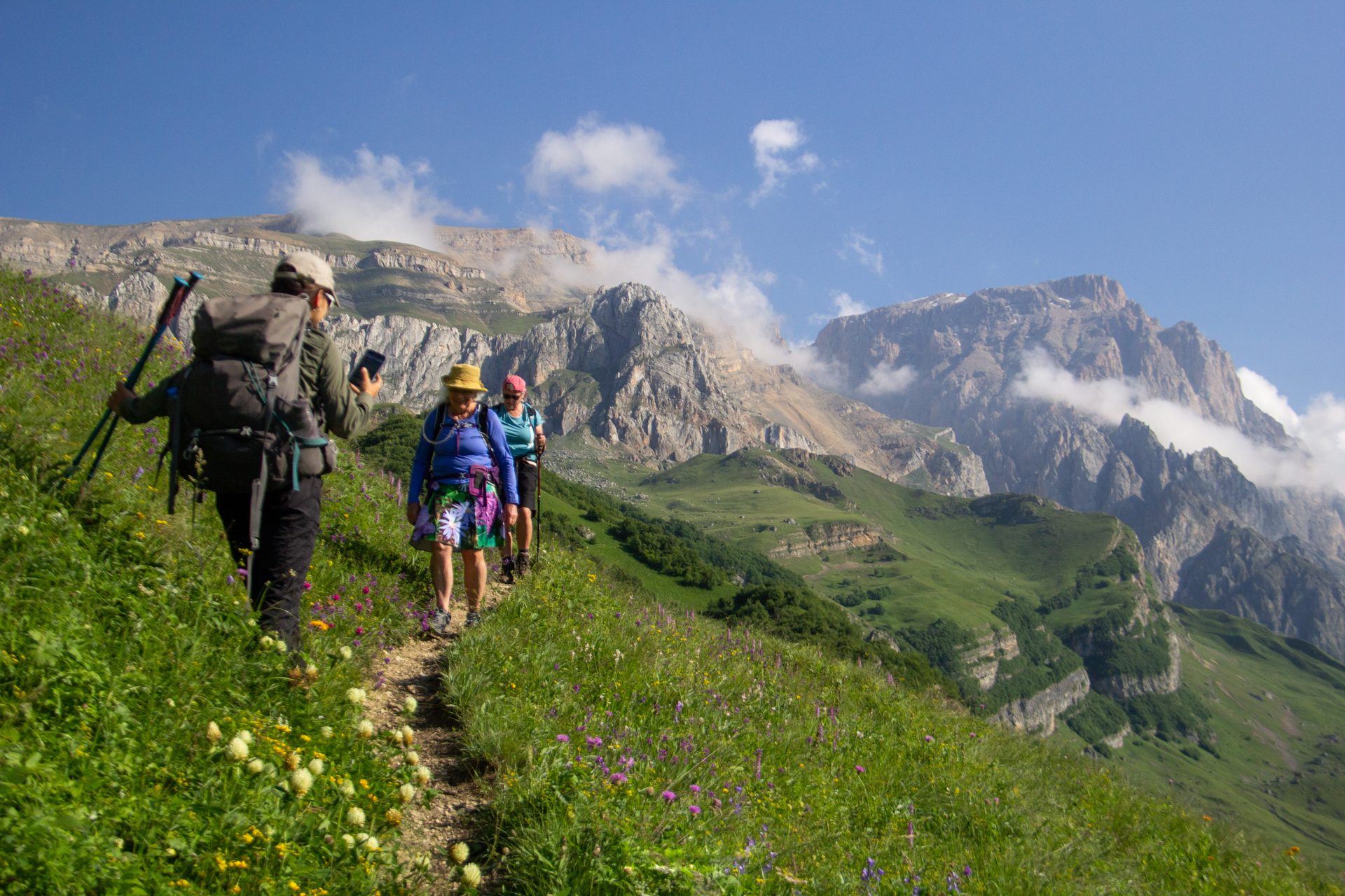 Three hikers on a narrow trail through a meadow full of wildflowers, with the limestone cliffs of Shahdag behind