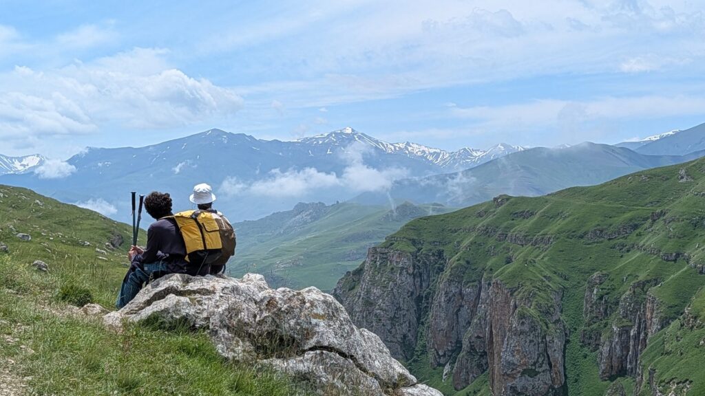 Two hikers resting on a rock above a deep green canyon, with snowy Caucasus peaks in the distance