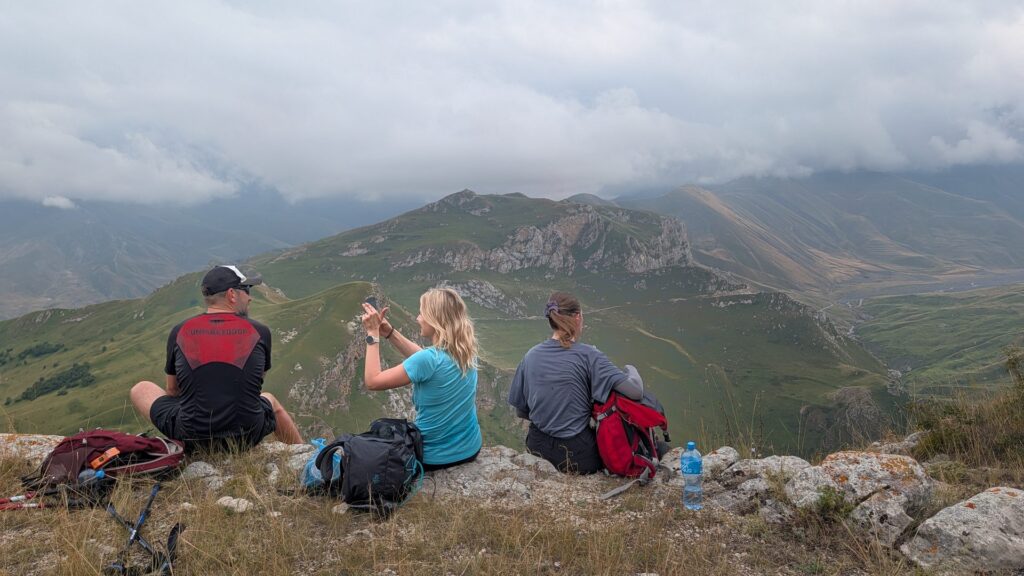 Three hikers sitting on a rocky ridge, looking over green mountain valleys under cloudy skies