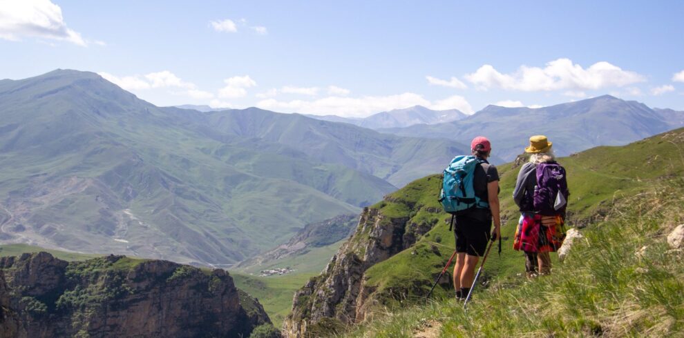 Two hikers on a grassy ridge above a deep canyon, with green mountains on the way from Khinalig towards Griz