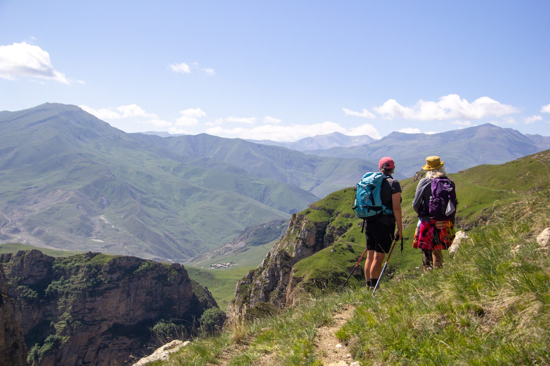Two hikers on a grassy ridge above a deep canyon, with green mountains on the way from Khinalig towards Griz