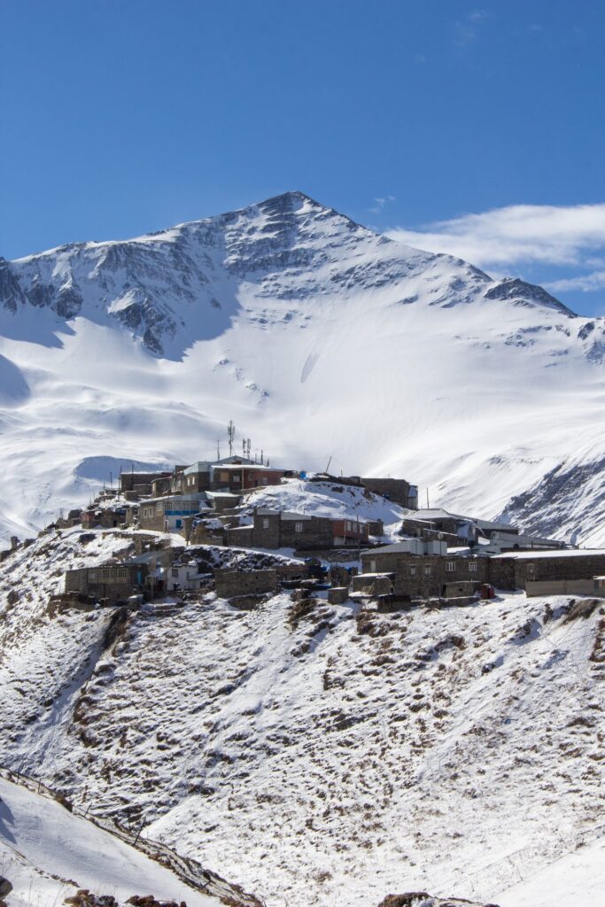 Khinalig village covered in snow on its hilltop, below a snowy peak and a deep blue sky